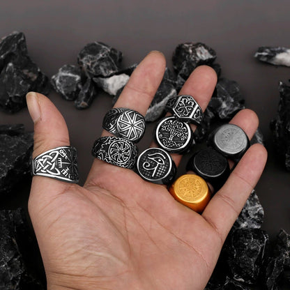 A hand displaying a selection of Viking and Norse mythology-inspired rings, featuring symbols like the Vegvísir, Tree of Life, Helm of Awe, and Egyptian Eye of Horus. The rings are crafted in black, silver, and gold-toned finishes, set against dark stones.