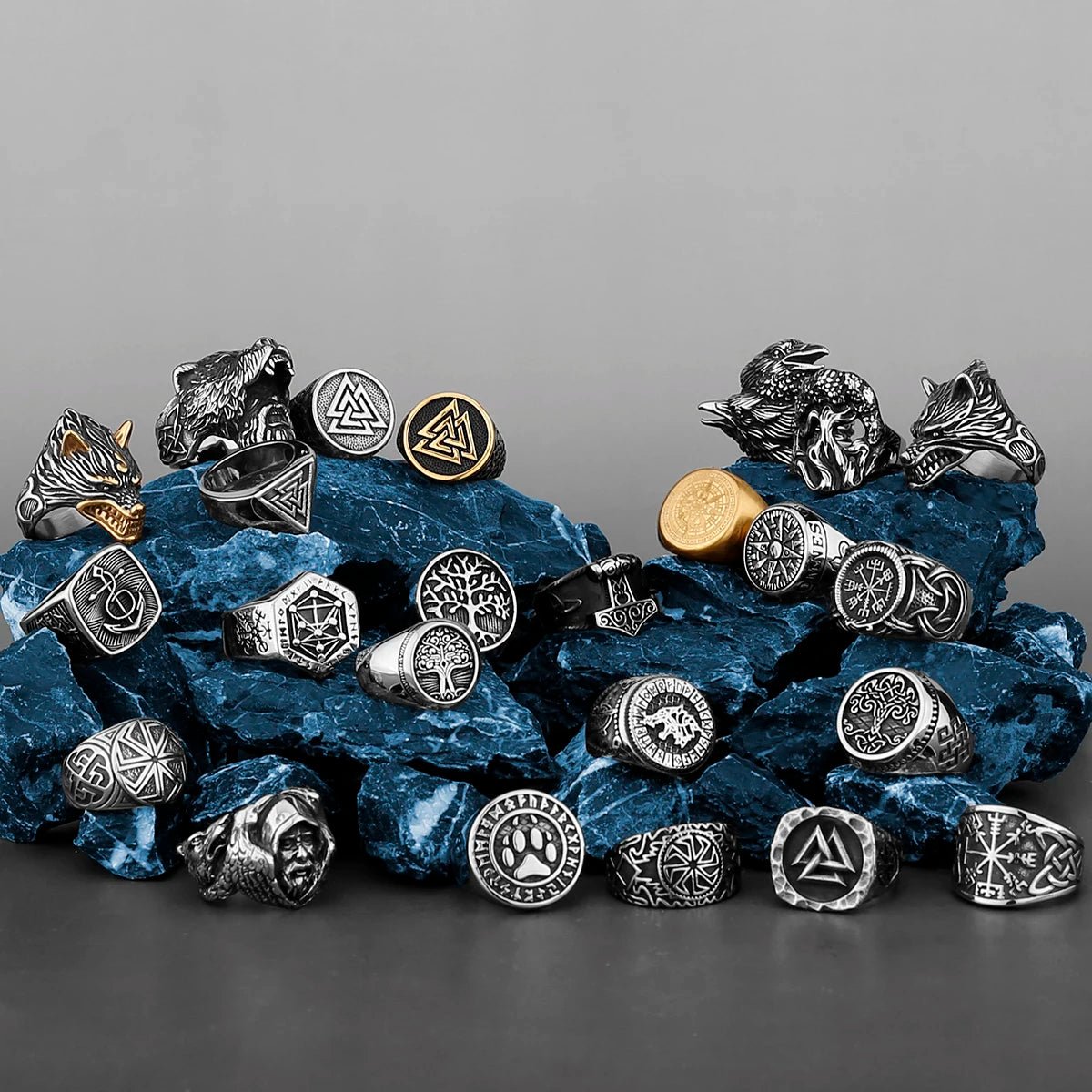 An artistic display of Viking-inspired rings arranged on textured blue stones. The collection features various Norse symbols, including the Valknut, Vegvísir, Yggdrasil, and Fenrir designs, in black, silver, and gold finishes.