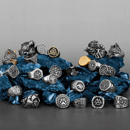 An artistic display of Viking-inspired rings arranged on textured blue stones. The collection features various Norse symbols, including the Valknut, Vegvísir, Yggdrasil, and Fenrir designs, in black, silver, and gold finishes.