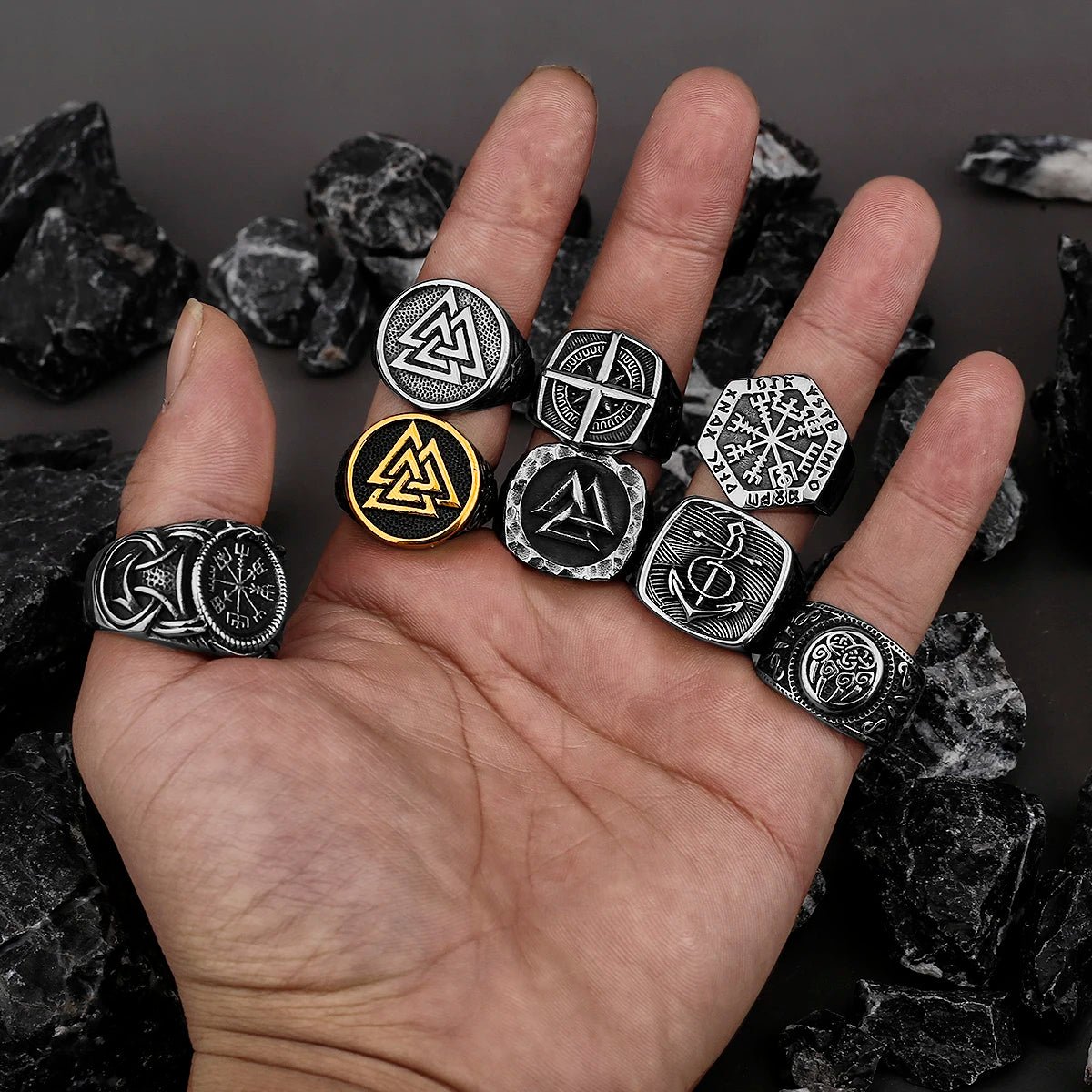 A hand displaying multiple Viking-inspired rings with intricate Norse symbols, including the Valknut, Vegvísir, Helm of Awe, and Yggdrasil. The rings come in black, silver, and gold finishes, set against a background of dark stones.